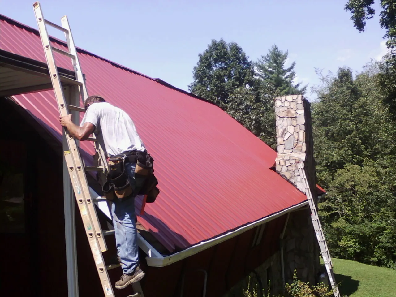 Roofer on a ladder beside a red metal roof and stone chimney — Soffit & Fascia Repair by Roofing Brothers in Deadwood